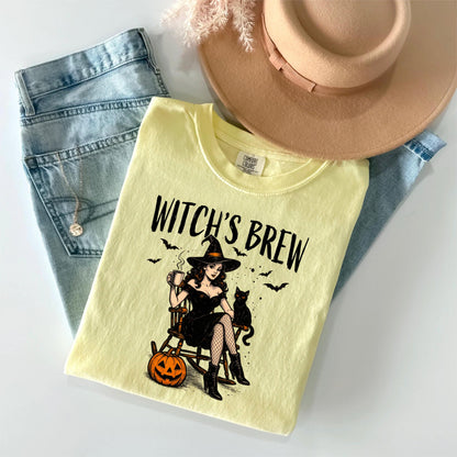 May include: A pale yellow t-shirt featuring a graphic of a person in a black dress and witch hat sitting in a rocking chair with a black cat, a pumpkin, and bats. The text "WITCH'S BREW" is printed above the illustration in a black font.