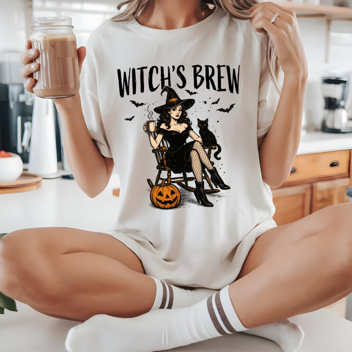 May include: An off-white t-shirt featuring a graphic print with the text WITCH'S BREW. The illustration depicts a person in a black dress and hat sitting in a rocking chair with a black cat, a pumpkin, and bats. The design is centered on the chest.