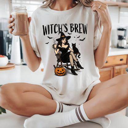 May include: An off-white t-shirt featuring a graphic print with the text WITCH'S BREW. The illustration depicts a person in a black dress and hat sitting in a rocking chair with a black cat, a pumpkin, and bats. The design is centered on the chest.