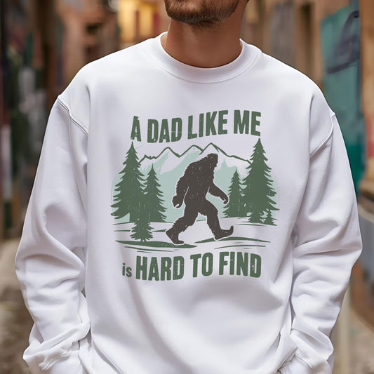 May include: White sweatshirt with a graphic featuring a Bigfoot silhouette walking in front of pine trees and mountains. The text reads "A DAD LIKE ME is HARD TO FIND" in green letters.