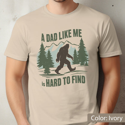 May include: Ivory-colored t-shirt featuring a graphic of a Bigfoot walking in front of pine trees and mountains. The text reads "A DAD LIKE ME is HARD TO FIND" in green and white. The shirt is made of a soft material.