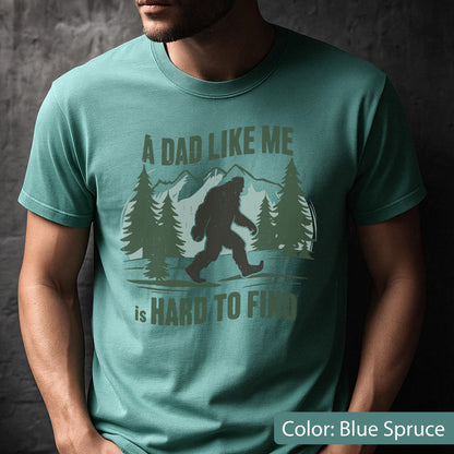 May include: Blue spruce t-shirt with a Bigfoot graphic and the text "A Dad Like Me is Hard to Find". The design includes a silhouette of Bigfoot walking in front of a mountain and trees.