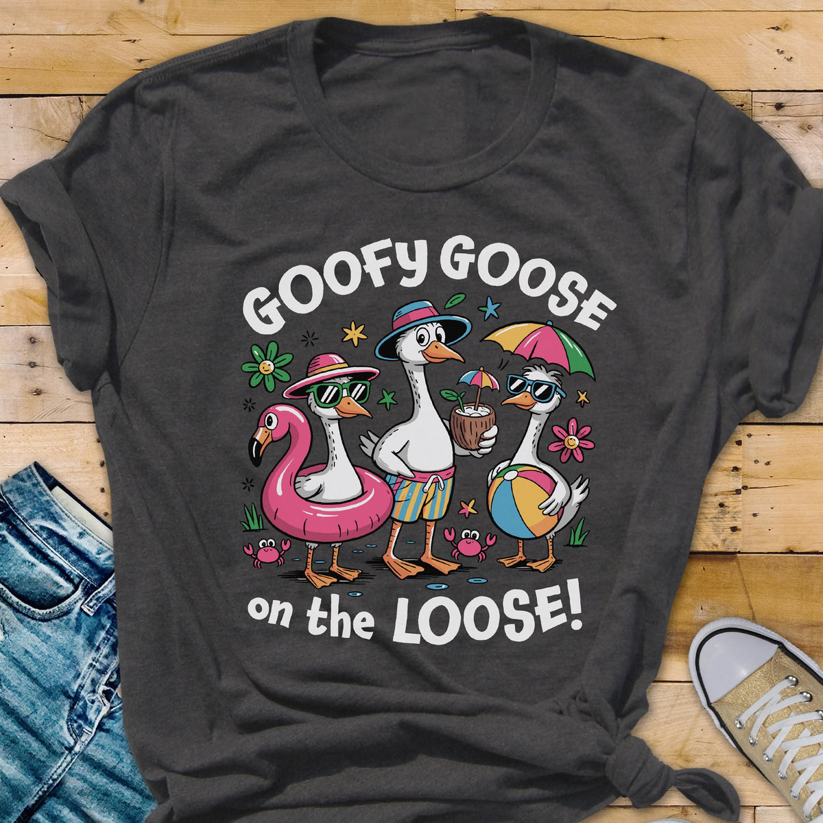 May include: Dark gray t-shirt featuring a cartoon design of three geese enjoying a beach day. The geese wear sunglasses, hats, and swimsuits, with a flamingo floatie, coconut drink, and beach ball. Text reads "GOOFY GOOSE on the LOOSE!"