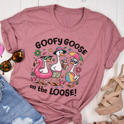 May include: A dusty rose t-shirt featuring a graphic of three cartoon geese in sunglasses and beach attire. The text "GOOFY GOOSE on the LOOSE!" is printed above and below the geese. The geese are on a beach scene with a flamingo floatie, a coconut drink, and a beach ball.