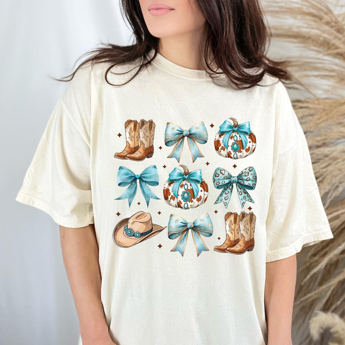 May include: Cream-colored t-shirt featuring a watercolor design of cowboy boots, bows, pumpkins, and a cowboy hat. The design incorporates shades of brown, blue, and white, with a rustic aesthetic. The shirt has a relaxed fit.