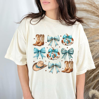 May include: Cream-colored t-shirt featuring a watercolor design of cowboy boots, bows, pumpkins, and a cowboy hat. The design incorporates shades of brown, blue, and white, with a rustic aesthetic. The shirt has a relaxed fit.