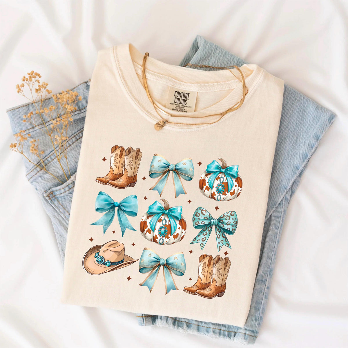 May include: Cream-colored t-shirt featuring a Western-themed design. The design includes illustrations of cowboy boots, bows, pumpkins, and a cowboy hat, all in shades of brown, turquoise, and white. The shirt is folded on top of a pair of blue jeans.