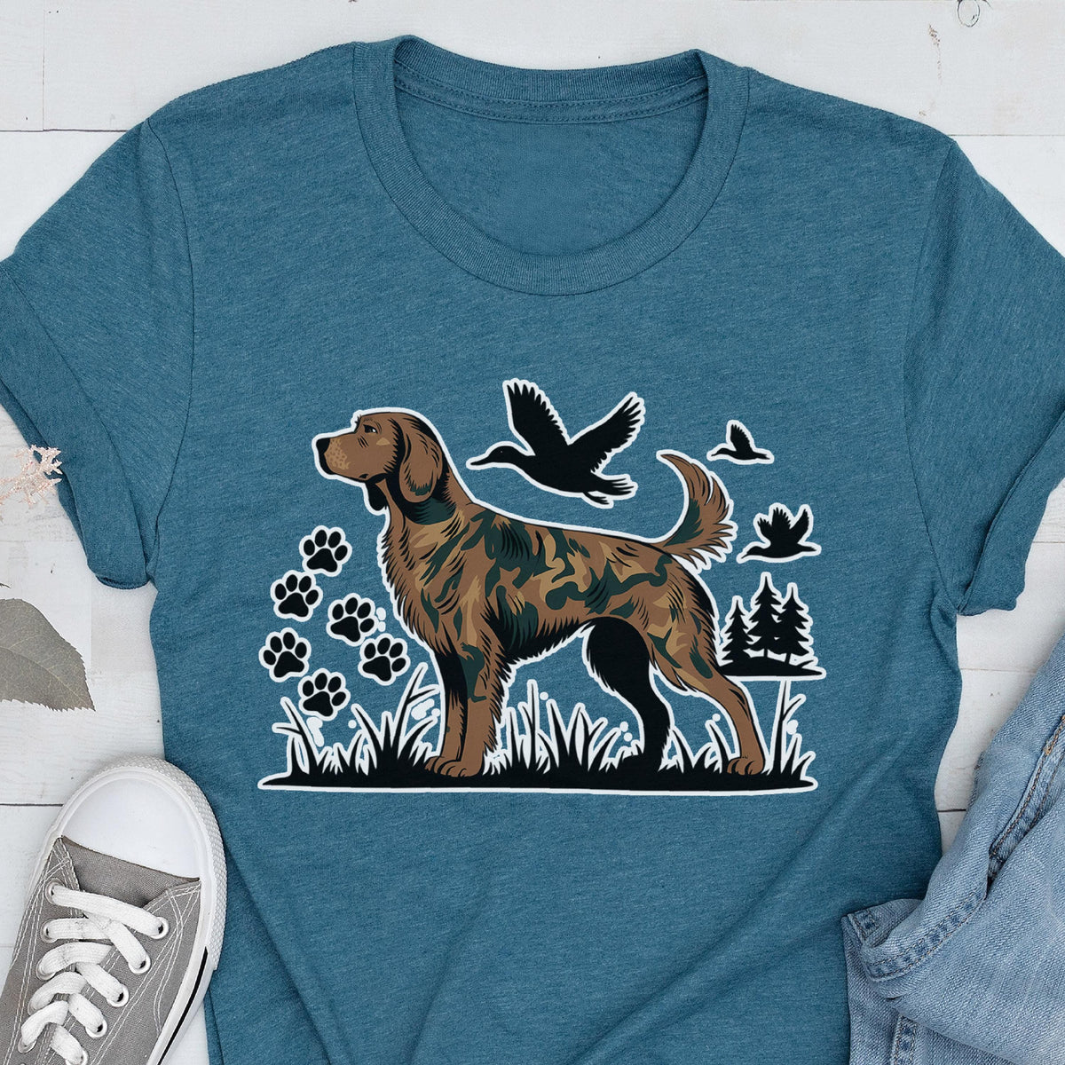 May include: A teal blue t-shirt featuring a graphic print of a brown dog with a camouflage pattern. The design includes black silhouettes of flying ducks, pine trees, and paw prints set against a backdrop of grass, creating a nature-themed scene.