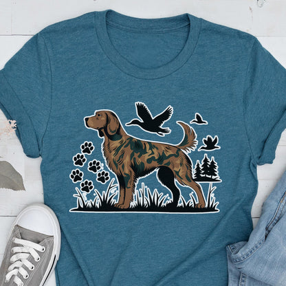 May include: A teal blue t-shirt featuring a graphic print of a brown dog with a camouflage pattern. The design includes black silhouettes of flying ducks, pine trees, and paw prints set against a backdrop of grass, creating a nature-themed scene.