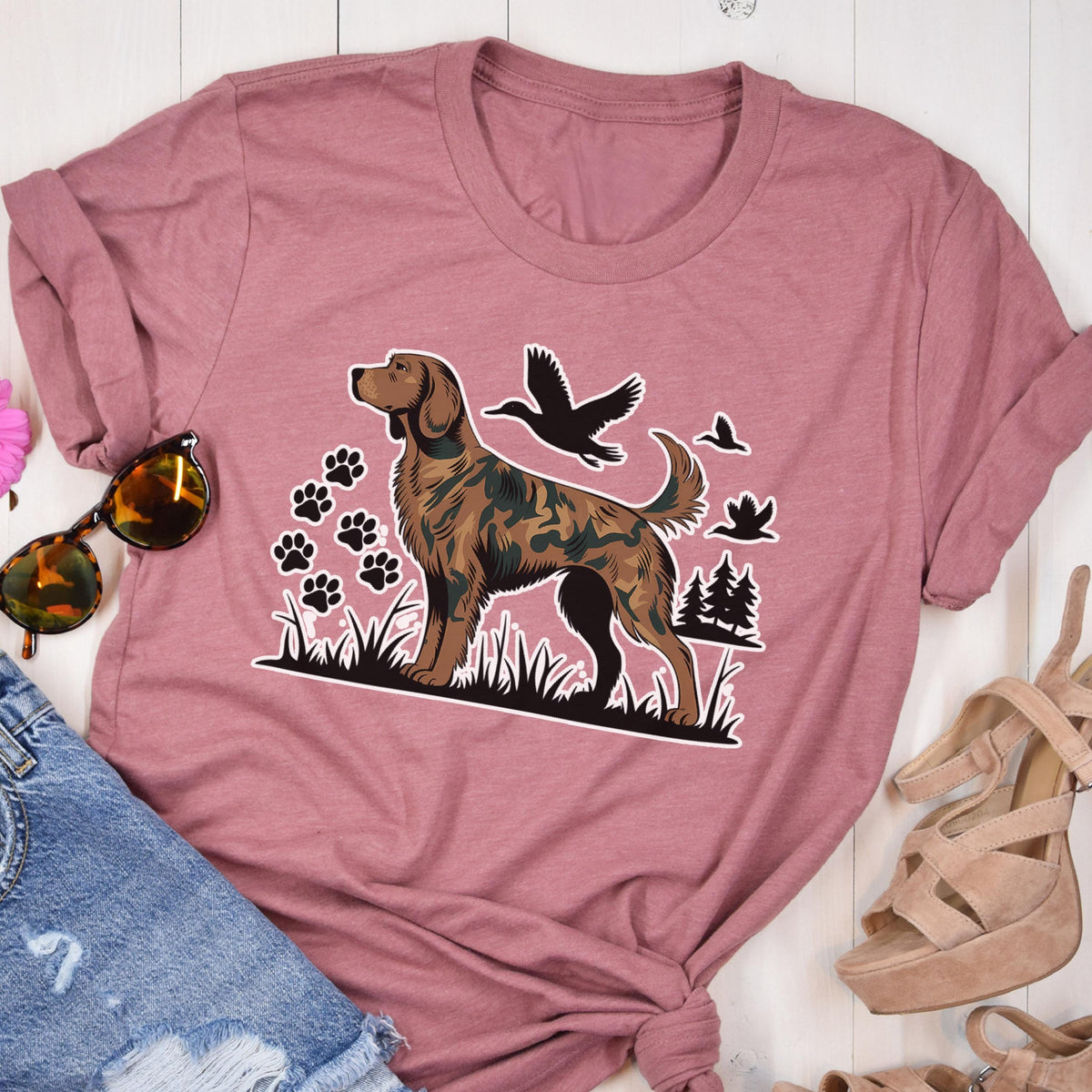 May include: A mauve crew neck t-shirt featuring a graphic print of a brown dog with a camouflage pattern on its body. The design includes black silhouettes of flying ducks, a small forest, and several paw prints set against a grassy base.