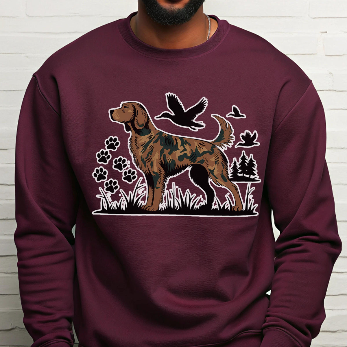 May include: Burgundy sweatshirt featuring a detailed illustration of a dog with a camouflage pattern. The design includes paw prints, birds, and trees. The dog is standing in grass, with a white outline around the entire design.