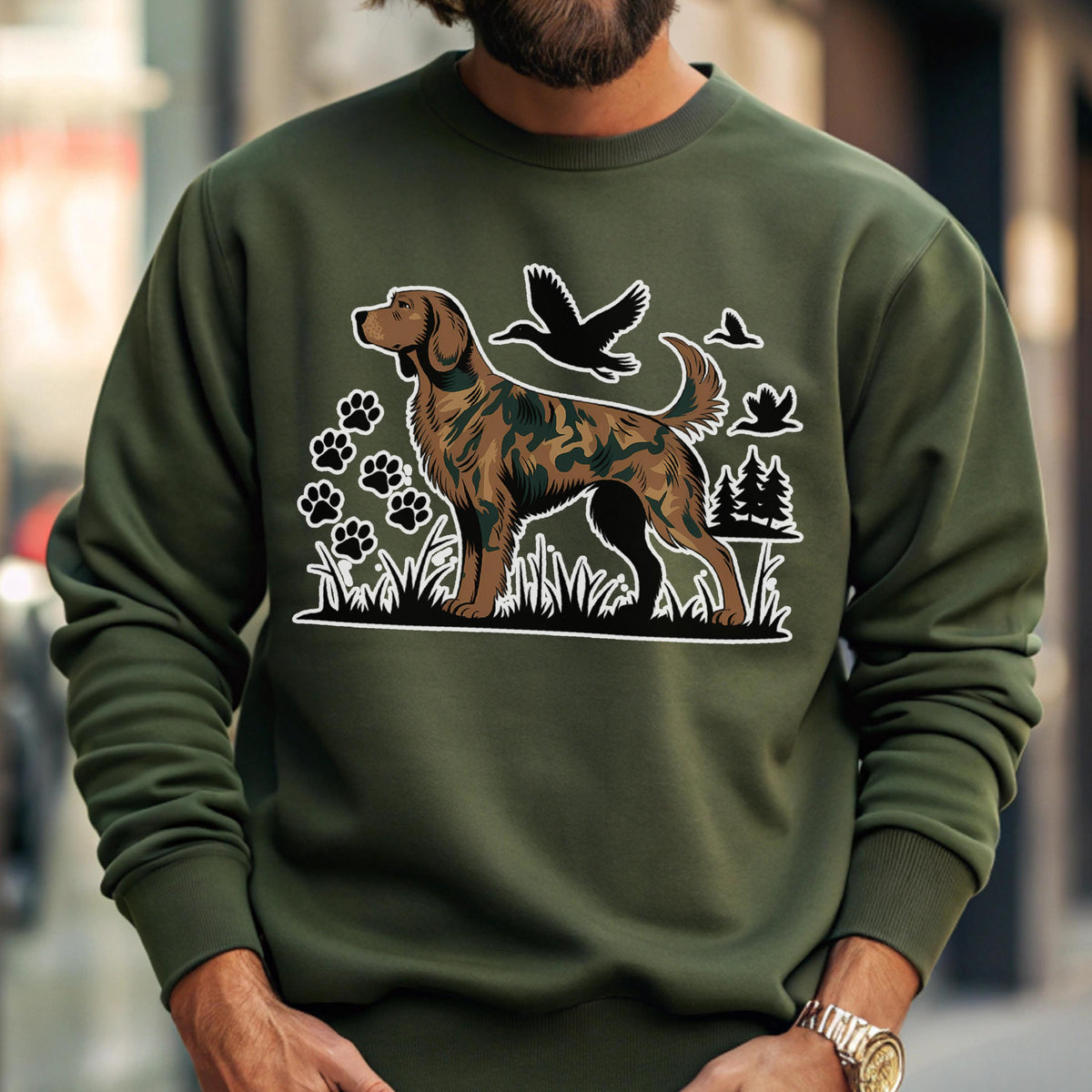 May include: Olive green sweatshirt featuring a graphic of a dog with a camouflage pattern, paw prints, and flying birds. The dog is brown and black, set against a white outline. The design includes grass and trees. The sweatshirt has ribbed cuffs and a crew neck.