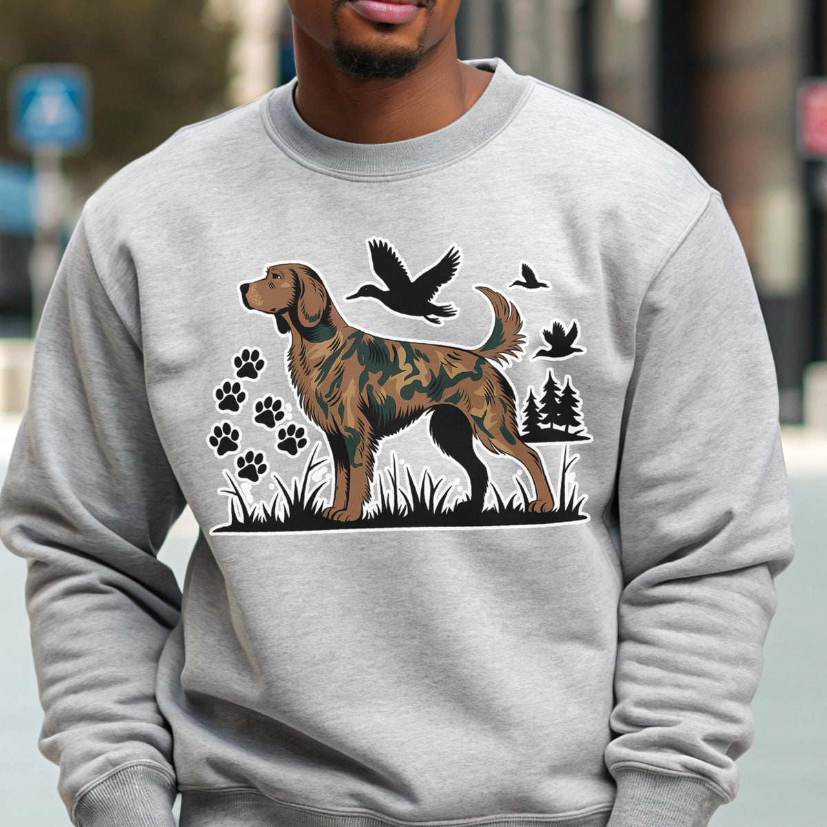 May include: A light gray sweatshirt featuring a brown and black dog illustration with a camouflage pattern. The design includes paw prints, flying birds, and trees. The sweatshirt is made of a soft material.
