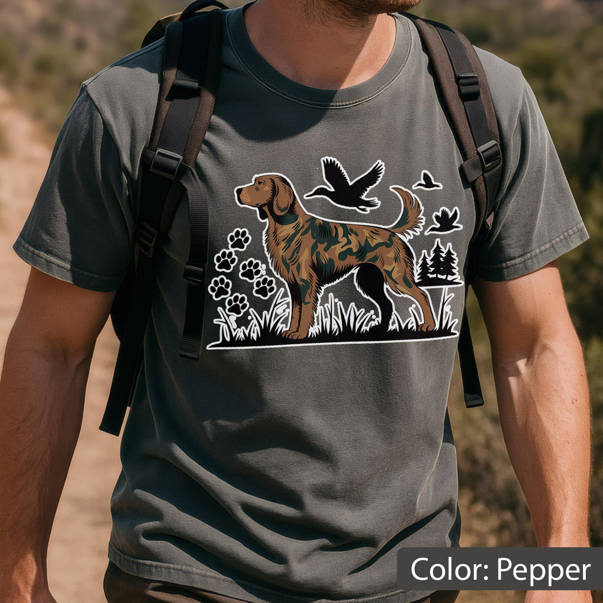 May include: A pepper-colored crew neck t-shirt featuring a graphic print of a dog with a camouflage pattern. The design includes silhouettes of flying ducks, pine trees, and paw prints arranged in a grassy field setting on the front chest.