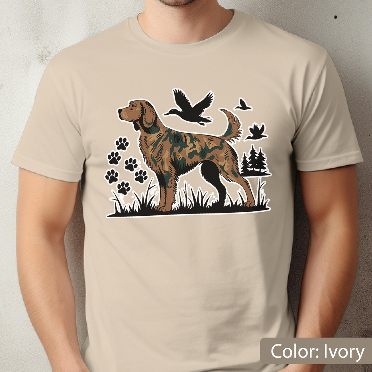 May include: An ivory t-shirt featuring a graphic print of a brown dog with a camouflage pattern. The design includes black silhouettes of flying ducks, pine trees, and several paw prints set against a backdrop of black grass.