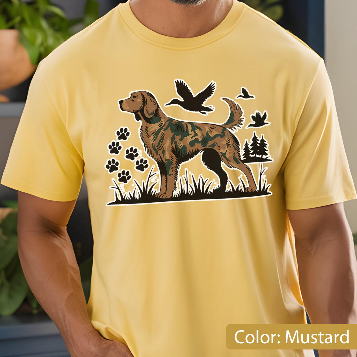 May include: A mustard yellow t-shirt featuring a graphic print of a dog with a camouflage pattern on its body. The design includes black silhouettes of flying ducks, paw prints, and pine trees set against a grassy landscape base.