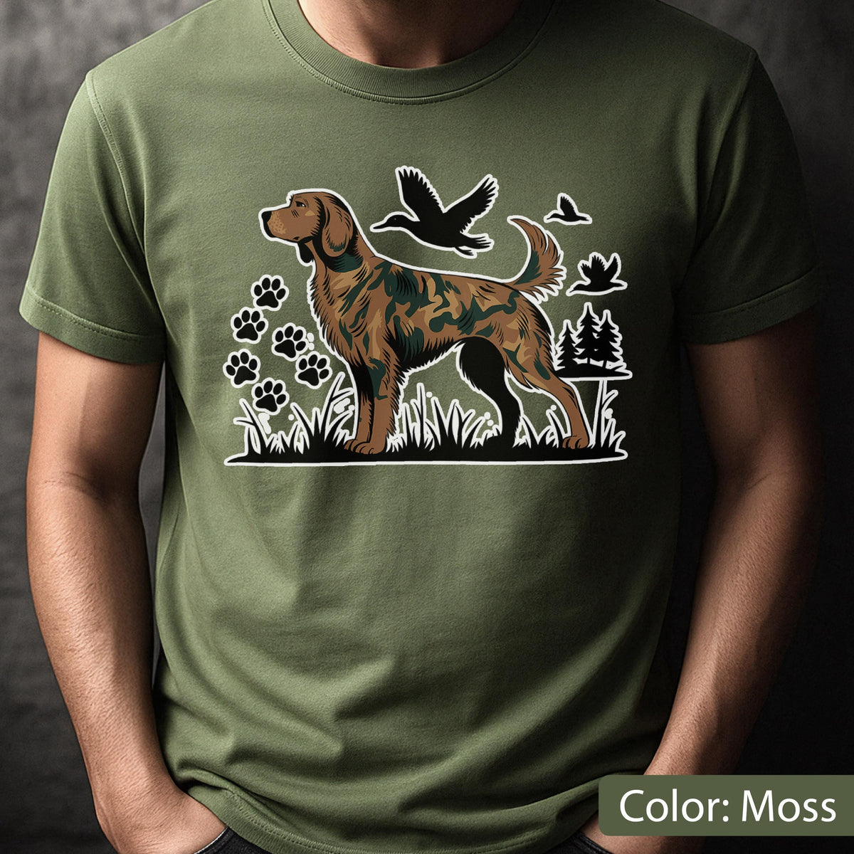 May include: A moss green short-sleeve t-shirt featuring a graphic print of a dog with a camouflage pattern. The design includes black silhouettes of flying ducks, pine trees, and several paw prints set against a grassy ground element.