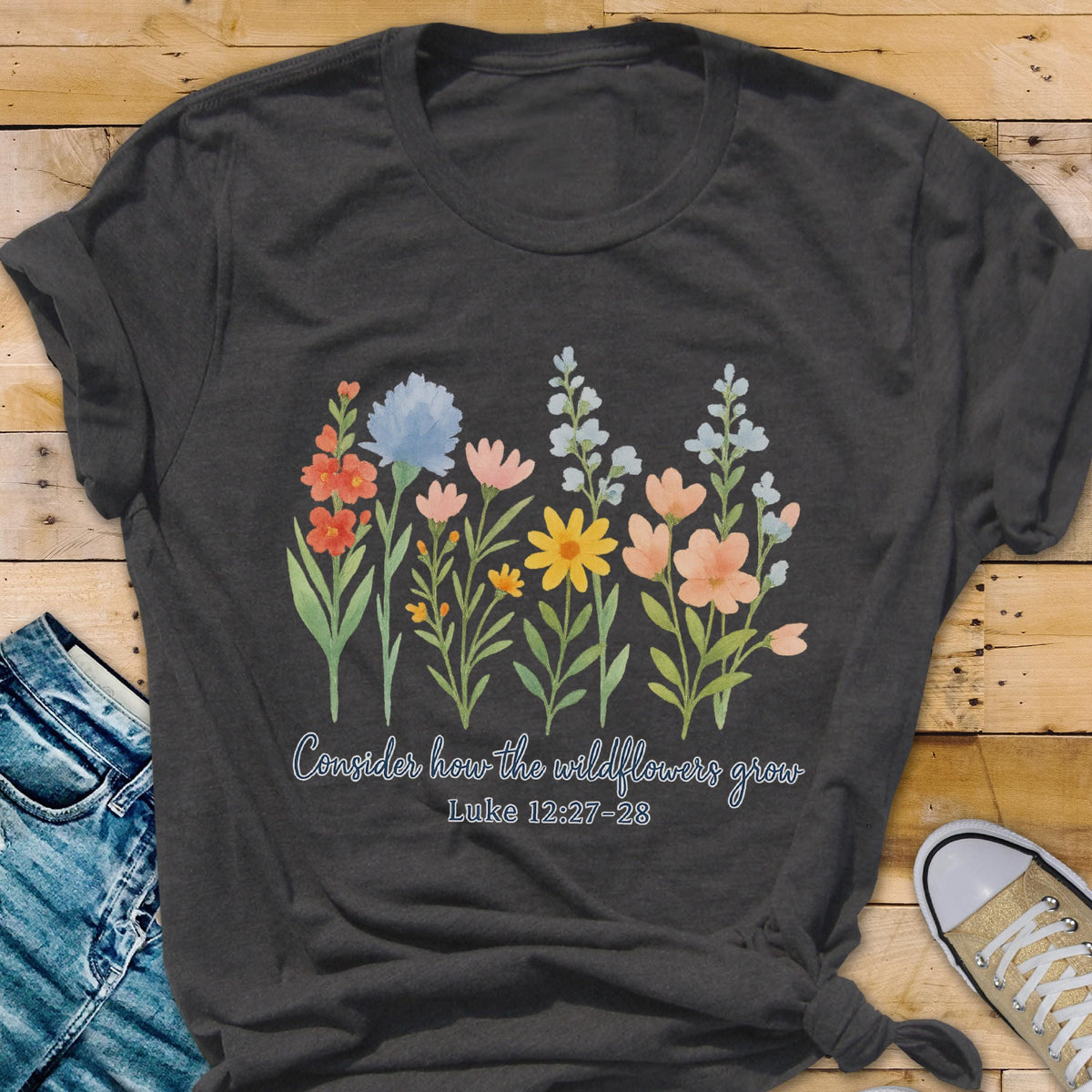 May include: Dark gray t-shirt featuring a watercolor floral design with orange, blue, yellow, and pink flowers. The text "Consider how the wildflowers grow" and "Luke 12:27-28" are printed below the flowers.