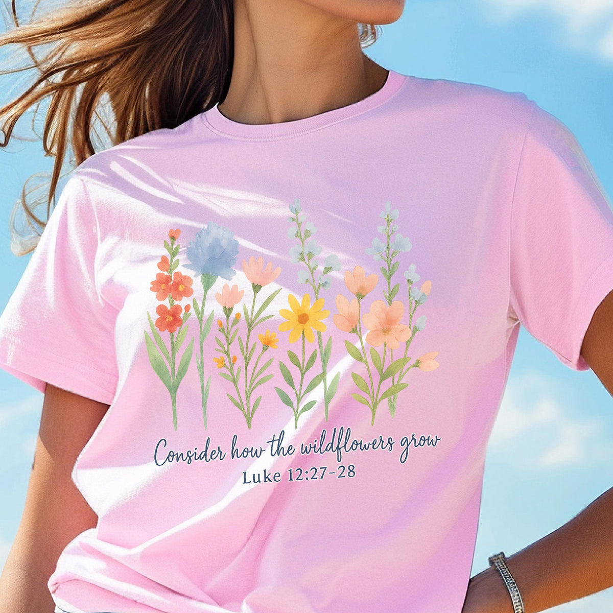 May include: A light pink t-shirt featuring a watercolor floral design with various wildflowers in shades of orange, yellow, blue, and peach. The text "Consider how the wildflowers grow, Luke 12:27-28" is printed below the flowers.