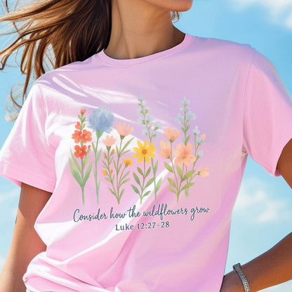May include: A light pink t-shirt featuring a watercolor floral design with various wildflowers in shades of orange, yellow, blue, and peach. The text "Consider how the wildflowers grow, Luke 12:27-28" is printed below the flowers.