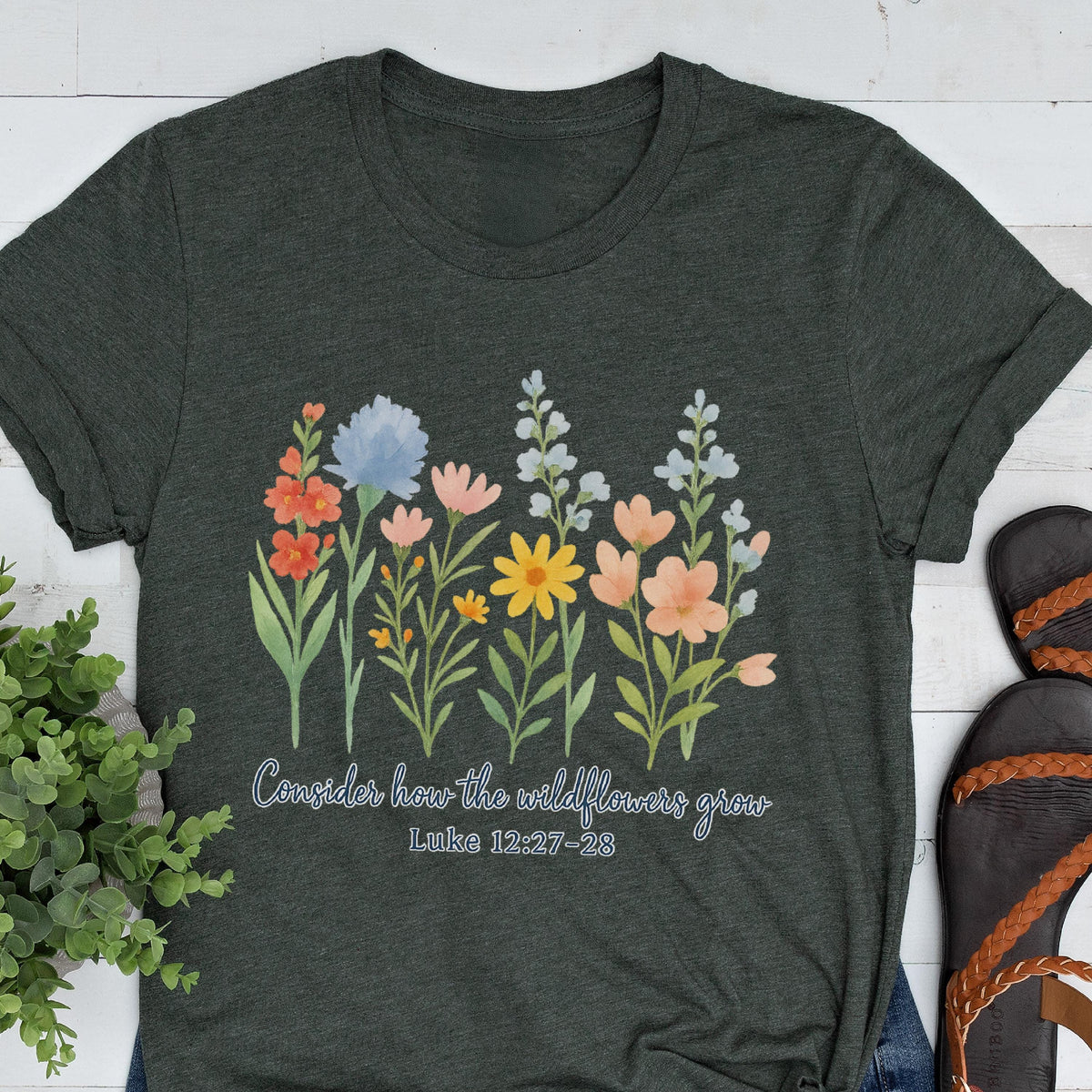 May include: Dark gray t-shirt featuring a watercolor floral design with orange, blue, pink, and yellow flowers. The text "Consider how the wildflowers grow" and Luke 12:27-28 are printed below the flowers.
