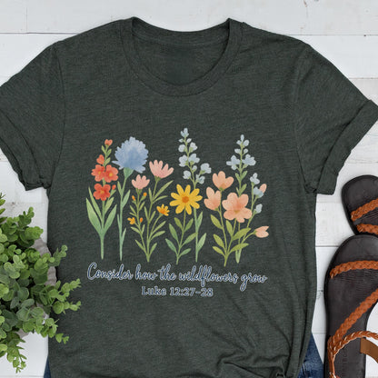 May include: Dark gray t-shirt featuring a watercolor floral design with orange, blue, pink, and yellow flowers. The text "Consider how the wildflowers grow" and Luke 12:27-28 are printed below the flowers.
