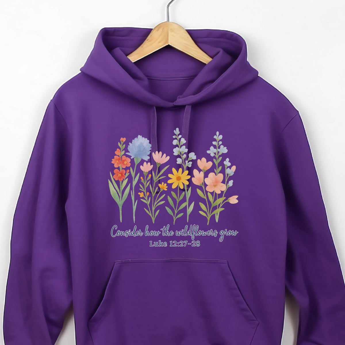 May include: A purple hoodie featuring a floral design with various wildflowers in shades of orange, blue, pink, and yellow. The text "Consider how the wildflowers grow" and Luke 12:27-28 are printed below the flowers.