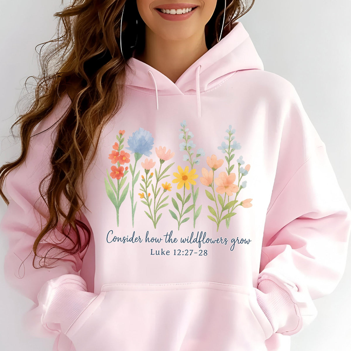 May include: A light pink hoodie featuring a watercolor floral design with various wildflowers in shades of orange, blue, yellow, and peach. The text "Consider how the wildflowers grow" and Bible verse Luke 12:27-28 are printed below the flowers.