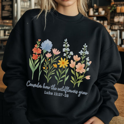 May include: Black sweatshirt featuring a watercolor floral design with various flowers in shades of orange, blue, pink, and yellow. The text "Consider how the wildflowers grow" and "Luke 12:27-28" are printed below the flowers.