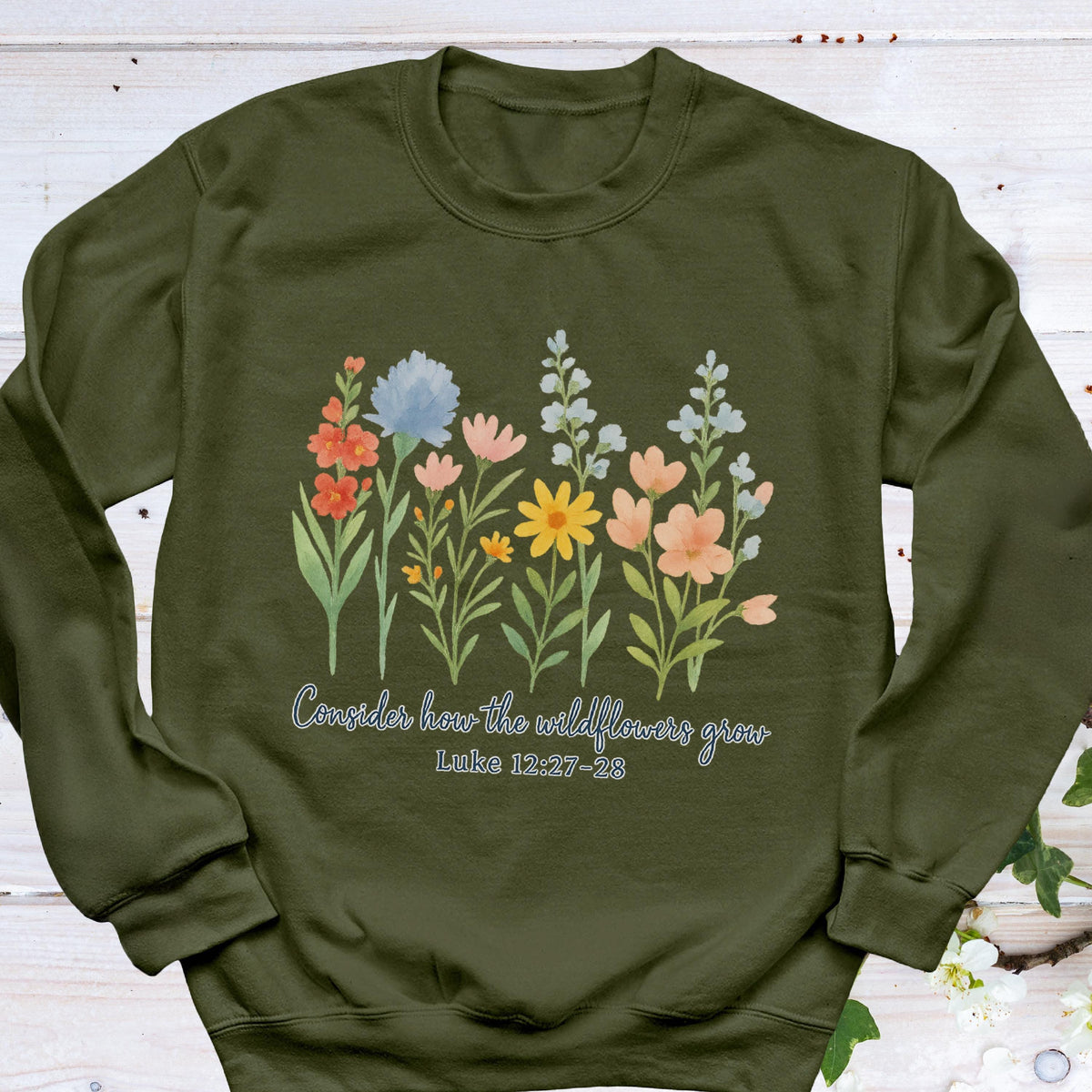 May include: Olive green sweatshirt featuring a watercolor floral design with various colorful wildflowers. The text "Consider how the wildflowers grow" and Luke 12:27-28 are printed below the flowers. The sweatshirt has a crew neck and long sleeves.