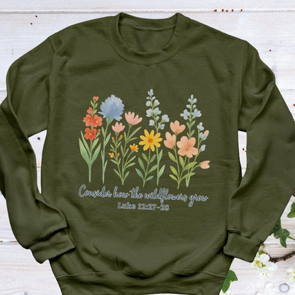 May include: Olive green sweatshirt featuring a watercolor floral design with various colorful wildflowers. The text "Consider how the wildflowers grow" and Luke 12:27-28 are printed below the flowers. The sweatshirt has a crew neck and long sleeves.