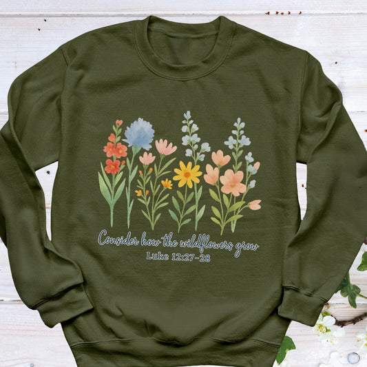 May include: Olive green sweatshirt featuring a watercolor floral design with various colorful wildflowers. The text "Consider how the wildflowers grow" and Luke 12:27-28 are printed below the flowers. The sweatshirt has a crew neck and long sleeves.