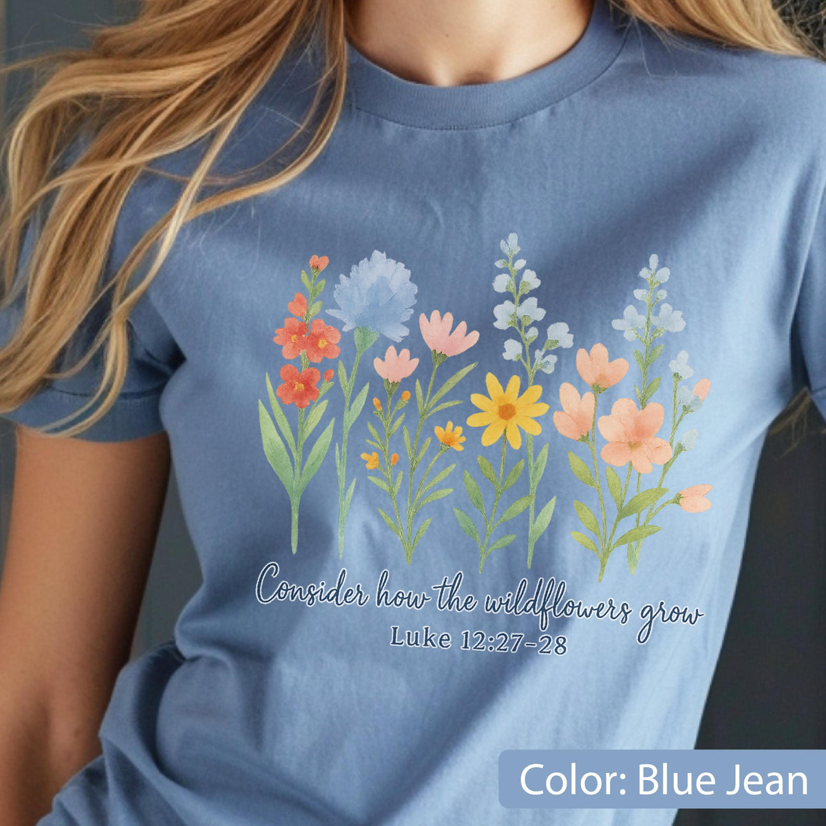 May include: A blue jean colored t-shirt featuring a watercolor-style graphic of various wildflowers in shades of orange, yellow, pink, and blue. Below the flowers, the text reads: Consider how the wildflowers grow Luke 12:27-28.