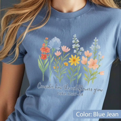 May include: A blue jean colored t-shirt featuring a watercolor-style graphic of various wildflowers in shades of orange, yellow, pink, and blue. Below the flowers, the text reads: Consider how the wildflowers grow Luke 12:27-28.