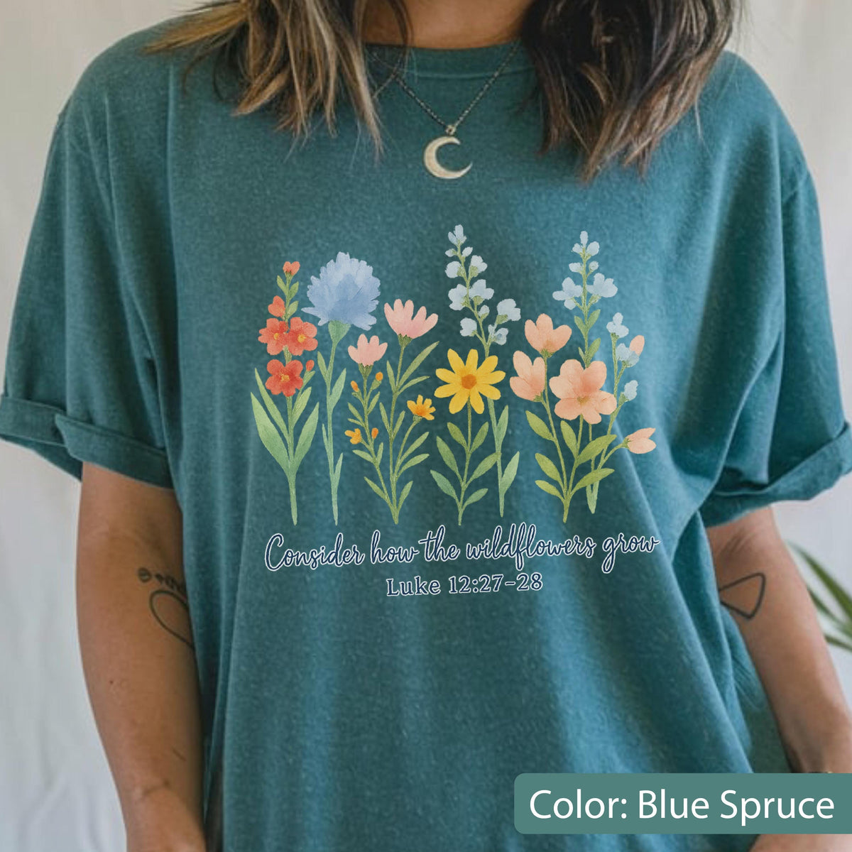May include: A blue spruce short-sleeve t-shirt featuring a colorful graphic of various wildflowers in shades of orange, yellow, blue, and pink. Below the flowers, the text reads, "Consider how the wildflowers grow" followed by "Luke 12:27-28".