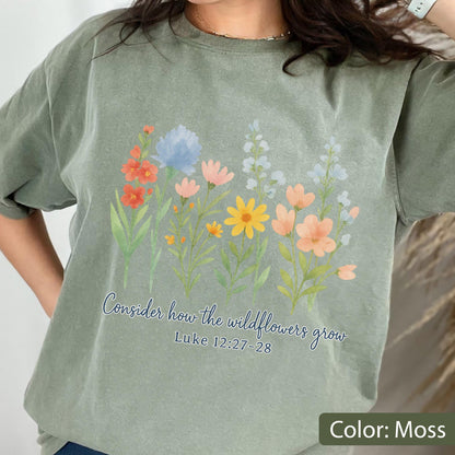 May include: A moss green short-sleeve t-shirt featuring a watercolor-style illustration of various wildflowers in shades of orange, yellow, and blue. Below the flowers, the text reads, "Consider how the wildflowers grow" and "Luke 12:27-28."