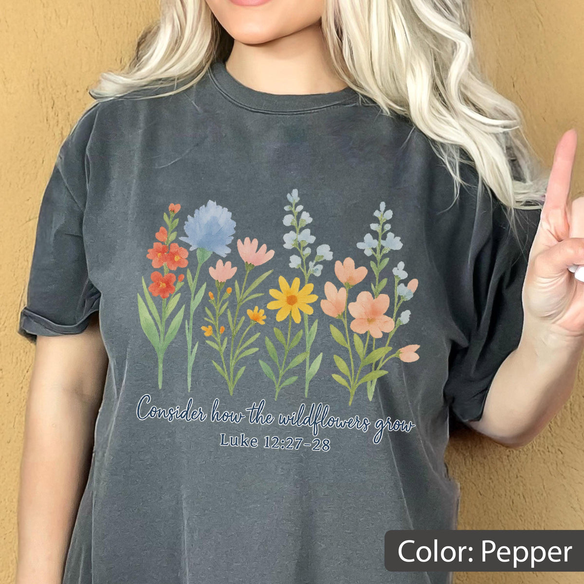 May include: A pepper-colored t-shirt featuring a colorful graphic of various wildflowers in shades of red, blue, pink, and yellow. Below the flowers, the text reads, "Consider how the wildflowers grow" followed by the scripture reference "Luke 12:27-28".