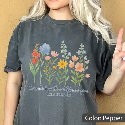 May include: A pepper-colored t-shirt featuring a colorful graphic of various wildflowers in shades of red, blue, pink, and yellow. Below the flowers, the text reads, "Consider how the wildflowers grow" followed by the scripture reference "Luke 12:27-28".