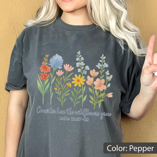 May include: A pepper-colored t-shirt featuring a colorful graphic of various wildflowers in shades of red, blue, pink, and yellow. Below the flowers, the text reads, "Consider how the wildflowers grow" followed by the scripture reference "Luke 12:27-28".