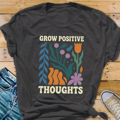 May include: Dark gray t-shirt with the words "GROW POSITIVE" and "THOUGHTS" in a cream-colored font. The shirt features a colorful floral design with orange, purple, teal, and blue elements. The shirt is made of a soft material.
