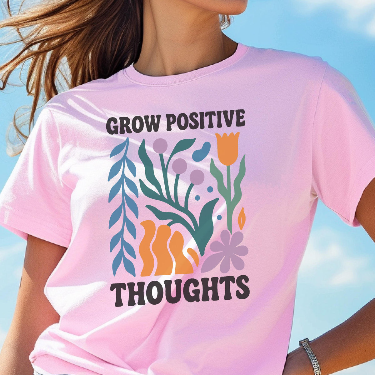 May include: A light pink t-shirt with the words "GROW POSITIVE THOUGHTS" in dark brown letters. The shirt features a colorful floral design with blue, green, orange, and purple elements. The shirt is made of a soft material.