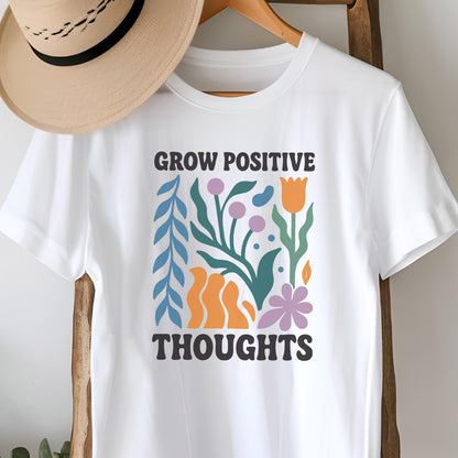 May include: White t-shirt with the text "GROW POSITIVE THOUGHTS" in black. The shirt features a colorful floral design with blue, orange, purple, and green elements. A tan hat hangs above the shirt on a wooden hanger.