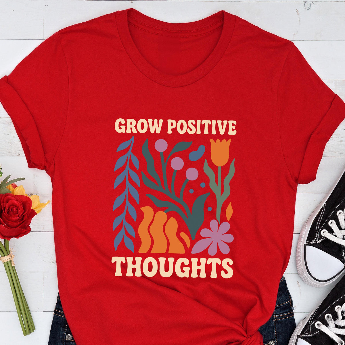 May include: A red t-shirt with the words "GROW POSITIVE THOUGHTS" in a cream-colored font. The shirt features a colorful floral design with blue, orange, purple, and green elements. Black sneakers are visible in the bottom right corner.