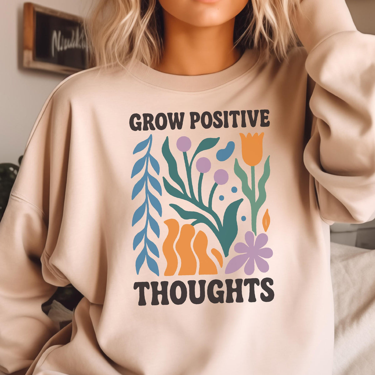 May include: A beige sweatshirt with the words "GROW POSITIVE THOUGHTS" in black block letters. The design includes colorful illustrations of flowers and leaves in blue, orange, purple, and green.