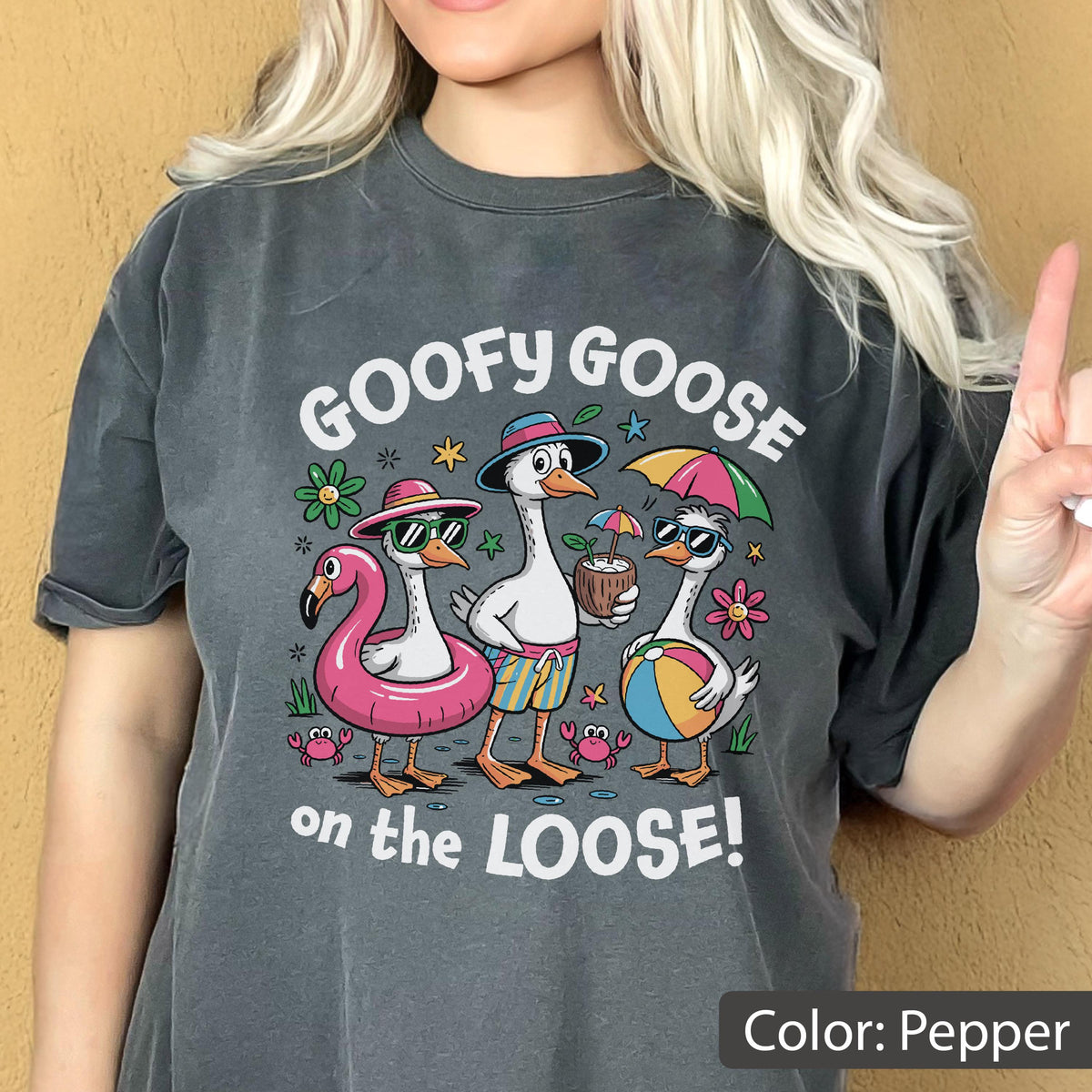 May include: A pepper-colored t-shirt featuring a graphic of three geese on vacation with the text GOOFY GOOSE on the LOOSE. The geese wear sunglasses, hats, and swim trunks, while interacting with a pink pool float, a coconut drink, and a beach ball.