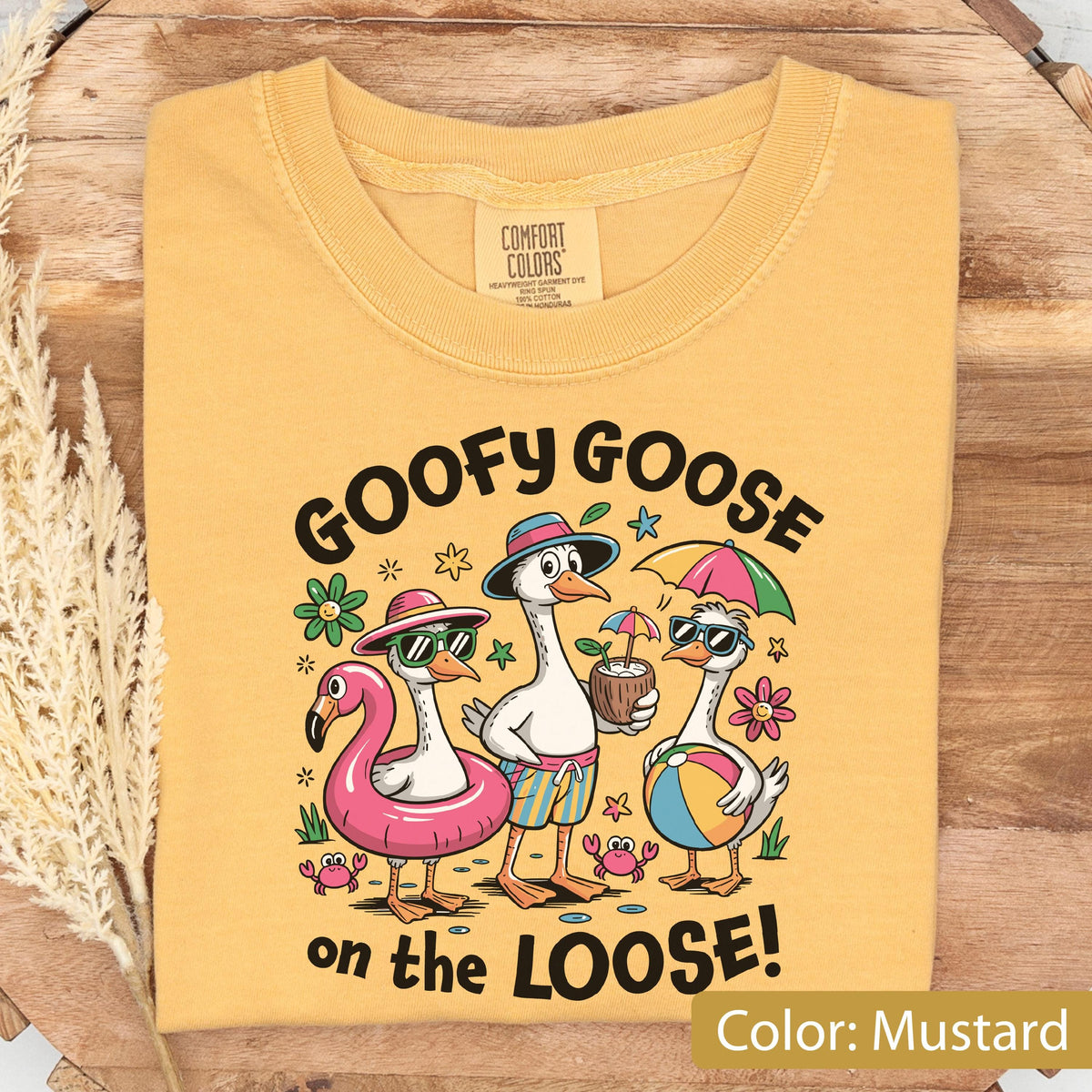 May include: A mustard yellow t-shirt featuring a graphic of three geese in summer attire with the text GOOFY GOOSE on the LOOSE. The geese are depicted with a pink flamingo pool float, a coconut drink, a beach ball, and colorful flowers.
