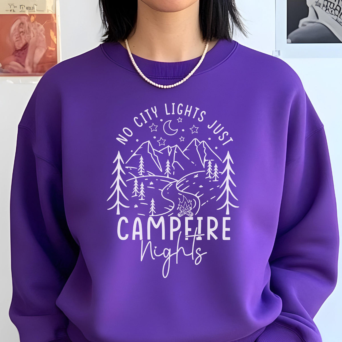 May include: A vibrant purple crewneck sweatshirt featuring a white graphic print of a mountain landscape, pine trees, a winding river, a campfire, and a crescent moon with stars. The text reads NO CITY LIGHTS JUST CAMPFIRE Nights in white.