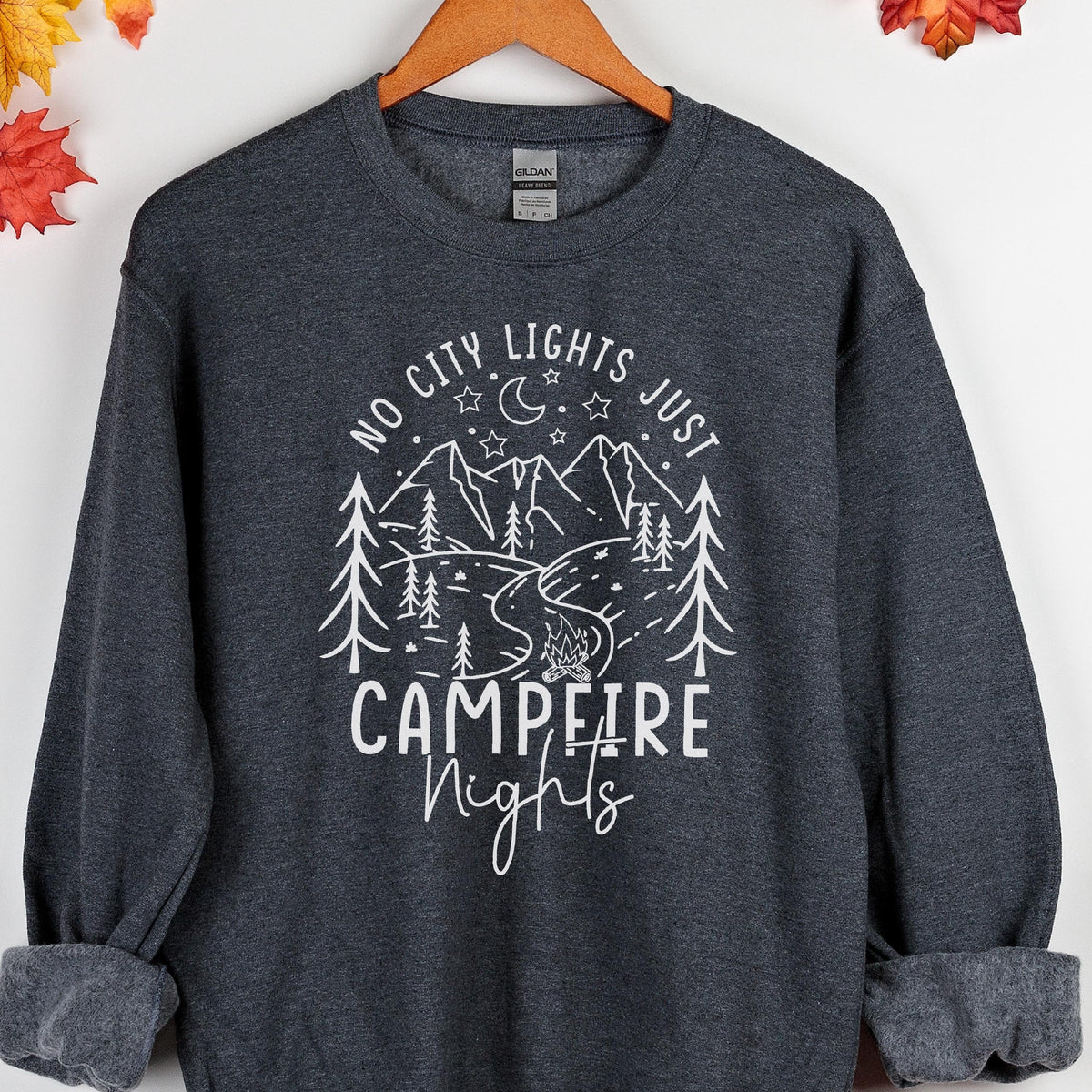 May include: A dark charcoal gray crewneck sweatshirt featuring a white graphic print of mountains, pine trees, a crescent moon, stars, and a campfire. The text reads "NO CITY LIGHTS JUST CAMPFIRE NIGHTS" in a mix of block and script lettering.