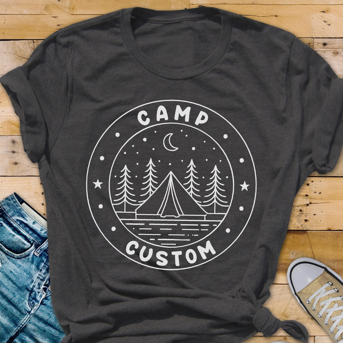 May include: A dark gray short-sleeve t-shirt featuring a white circular graphic. The design includes a tent, pine trees, a crescent moon, and stars, with the text CAMP at the top and CUSTOM at the bottom, all enclosed within a circular border.