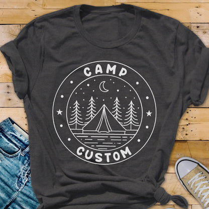 May include: A dark gray short-sleeve t-shirt featuring a white circular graphic. The design includes a tent, pine trees, a crescent moon, and stars, with the text CAMP at the top and CUSTOM at the bottom, all enclosed within a circular border.